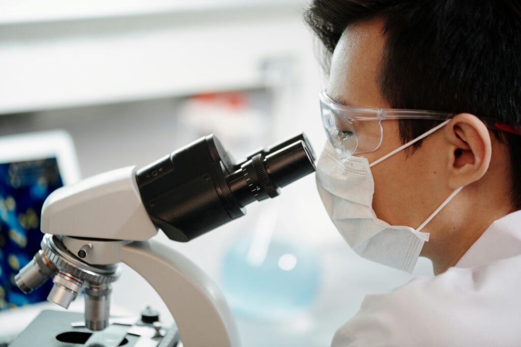 Scientist wearing face mask and goggles examining samples through a microscope in a lab.